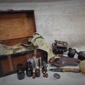 Op de afbeelding: Een houten schatkist met een messing slot en scharnieren, open om een ​​groene stoffen voering te onthullen. De kist is gevuld met verschillende items, waaronder een lederen dagboek, een kompas, een kolf en verschillende kleine flesjes. Er is ook een vintage camera, een verrekijker en een globe.