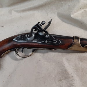 Op de afbeelding: Een antiek-stijl flintlock pistool met een houten handvat. Het pistool is zwart en messing, met een messing trekkerbeschermer en een messing loopband.