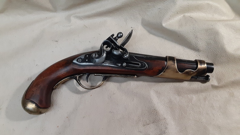 Op de afbeelding: Een antiek-stijl flintlock pistool met een houten handvat. Het pistool is zwart en messing, met een messing trekkerbeschermer en een messing loopband.