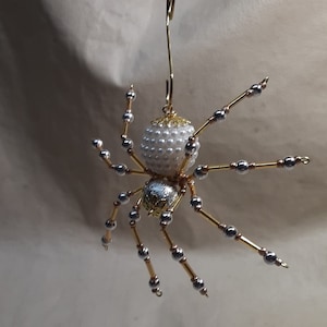 May include: A decorative spider ornament with a white pearl bead body and silver and gold accents. The spider's legs are made of gold wire with silver beads. The ornament is suspended by a gold wire hook.