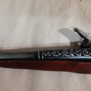 May include: A black and silver flintlock pistol with a wooden stock and intricate floral engravings. The hammer is cocked and the trigger is visible.