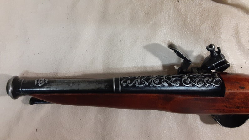May include: A black and silver flintlock pistol with a wooden stock and intricate floral engravings. The hammer is cocked and the trigger is visible.