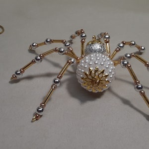 May include: A decorative spider ornament with a body covered in white beads and a gold and silver design. The legs are gold with silver and copper accents. A gold wire spiral is in the background.