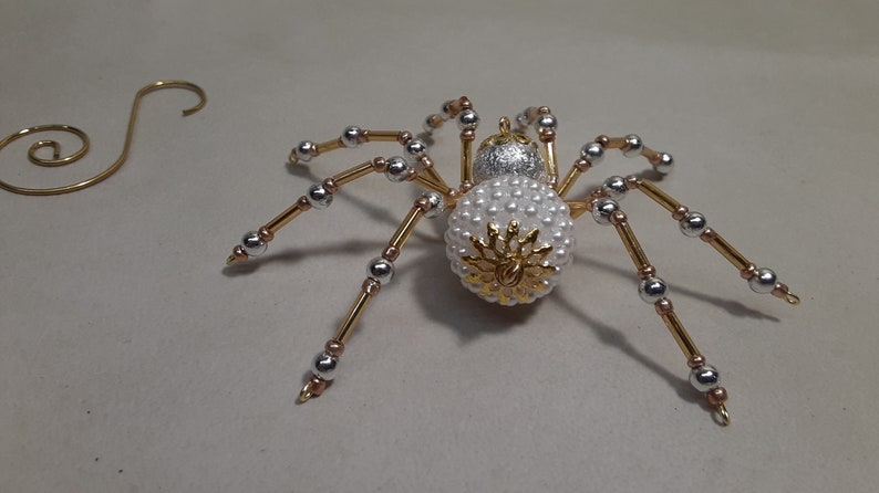 May include: A decorative spider ornament with a body covered in white beads and a gold and silver design. The legs are gold with silver and copper accents. A gold wire spiral is in the background.