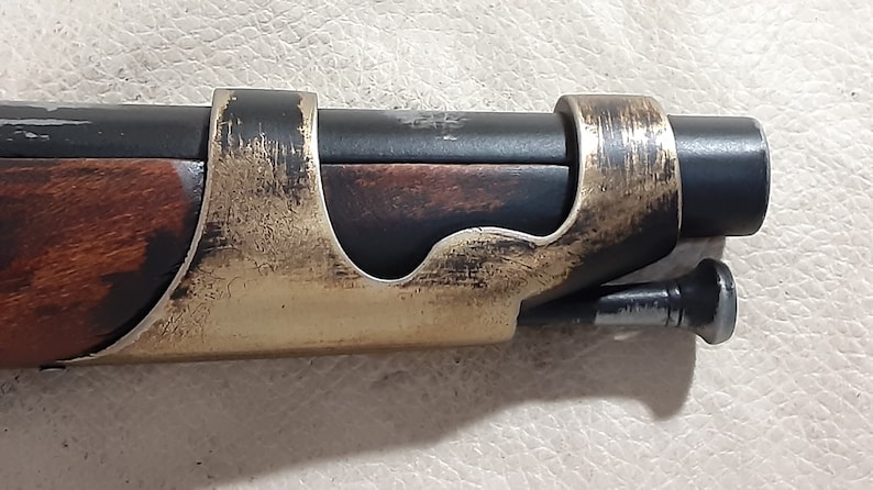 Op de afbeelding: Een close-up van een zwart en bruin houten pistool met een gouden metalen accent. Het pistool heeft een zwarte metalen loop en een houten handvat.