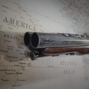 May include: A silver and brown double-barreled pistol with a wooden handle. The pistol is lying on a map of South America.