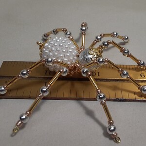 May include: A decorative spider ornament with a body made of white pearls and a silver and gold head. The legs are gold and silver, and the spider is approximately 3 inches long.