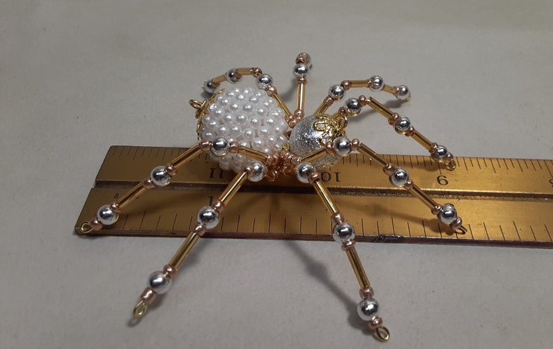 May include: A decorative spider ornament with a body made of white pearls and a silver and gold head. The legs are gold and silver, and the spider is approximately 3 inches long.
