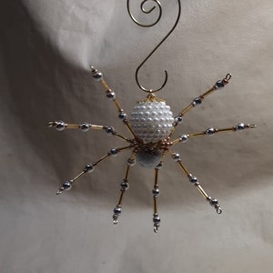 May include: A handmade spider ornament with a white pearl bead body and silver bead legs. The spider's legs are attached to a gold wire frame. A gold wire loop is at the top for hanging. This decorative item is perfect for Halloween or a unique gift.