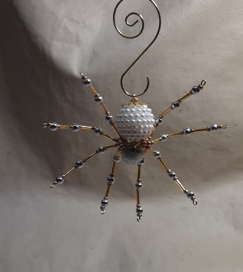 May include: A handmade spider ornament with a white pearl bead body and silver bead legs. The spider's legs are attached to a gold wire frame. A gold wire loop is at the top for hanging. This decorative item is perfect for Halloween or a unique gift.