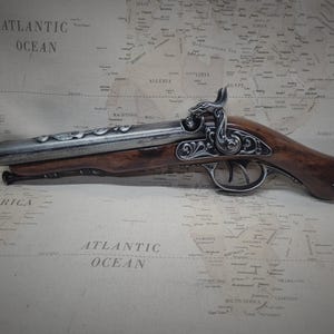 May include: A silver and brown pirate pistol with intricate details and a skull and crossbones on the handle. The pistol is lying on a map with the words "Atlantic Ocean" printed on it.