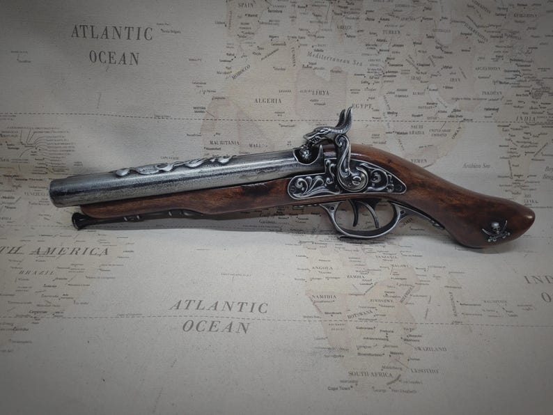 May include: A silver and brown pirate pistol with intricate details and a skull and crossbones on the handle. The pistol is lying on a map with the words "Atlantic Ocean" printed on it.