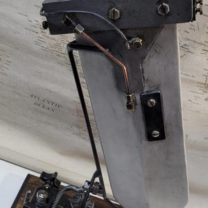 Può includere: Una scultura in metallo in stile steampunk con una parte superiore avvolta in pelle marrone scuro, un corpo grigio e una base in legno. La scultura presenta vari componenti meccanici, tra cui ingranaggi, quadranti e tubi di rame. Lo sfondo include una mappa vintage.