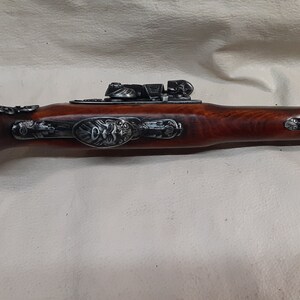 May include: A black and silver flintlock pistol with a wooden stock. The pistol has intricate carvings and a decorative metal plate on the side.