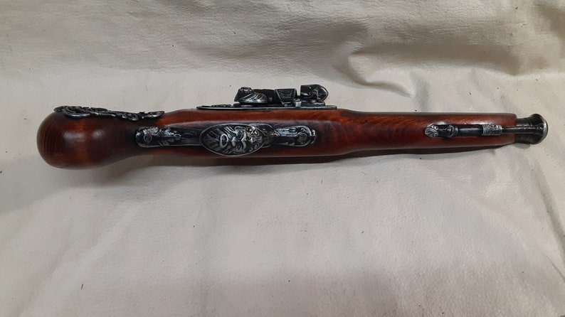 May include: A black and silver flintlock pistol with a wooden stock. The pistol has intricate carvings and a decorative metal plate on the side.