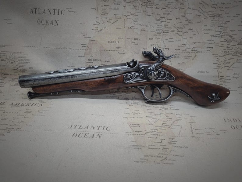May include: A silver and brown pirate pistol with intricate details and a skull and crossbones design on the handle. The pistol is lying on a vintage world map.