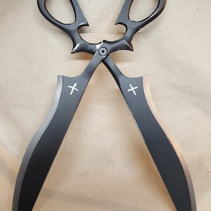 May include: Black decorative scissors with a cross design on each blade. The scissors have a unique, ornate design and are made of metal.