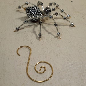 May include: A silver and black beaded spider ornament with a gold hook. The spider has a detailed, patterned body and a clear, faceted gem in the center.