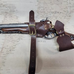 May include: A brown leather holster with a metal buckle and a brass-coloured trigger guard. The holster is attached to a brown leather belt with a metal buckle. A black and silver pistol is inside the holster.