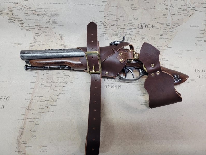 May include: A brown leather holster with a metal buckle and a brass-coloured trigger guard. The holster is attached to a brown leather belt with a metal buckle. A black and silver pistol is inside the holster.