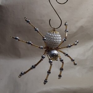 May include: A handcrafted spider ornament with a white beaded body and silver and gold legs. The spider has a decorative gold head and a gold wire loop for hanging. This unique ornament is perfect for Halloween or year-round decor.