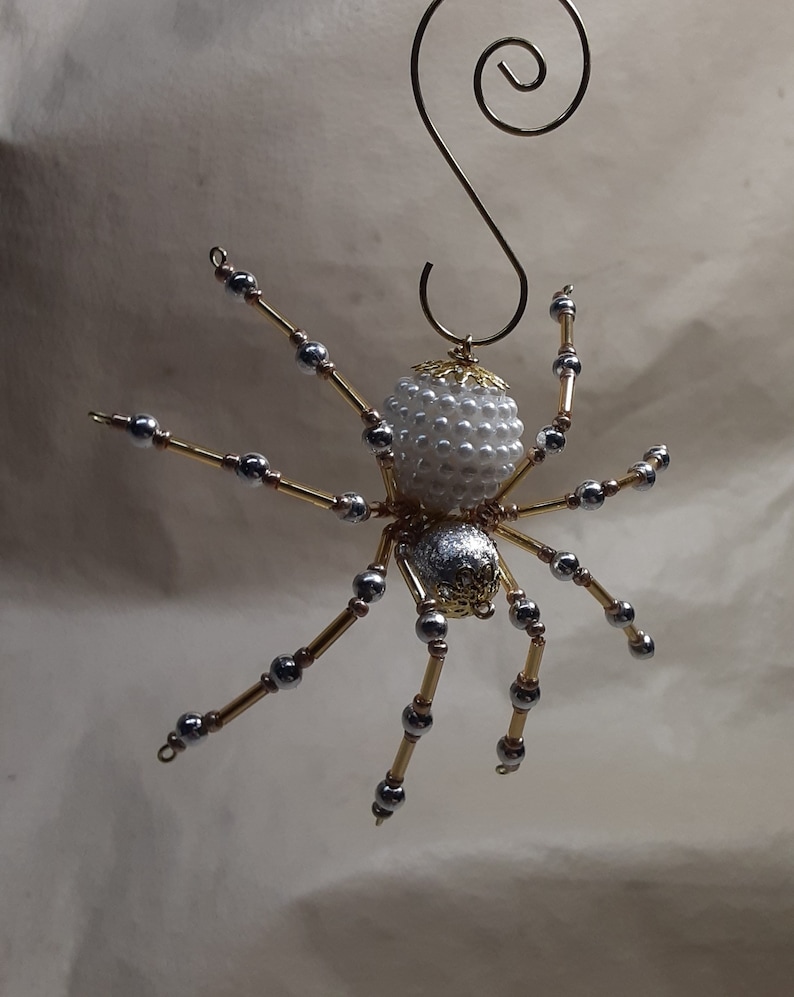 May include: A handcrafted spider ornament with a white beaded body and silver and gold legs. The spider has a decorative gold head and a gold wire loop for hanging. This unique ornament is perfect for Halloween or year-round decor.