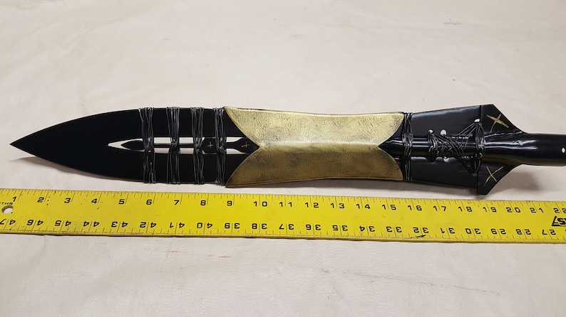 May include: A black and gold spearhead with a leather wrap. The spearhead is about 20 inches long and is measured against a ruler.