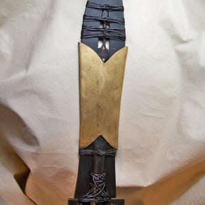 May include: A black and gold spear with a pointed tip and a decorative metal band around the middle. The spear is wrapped in black cord and has a wooden handle.