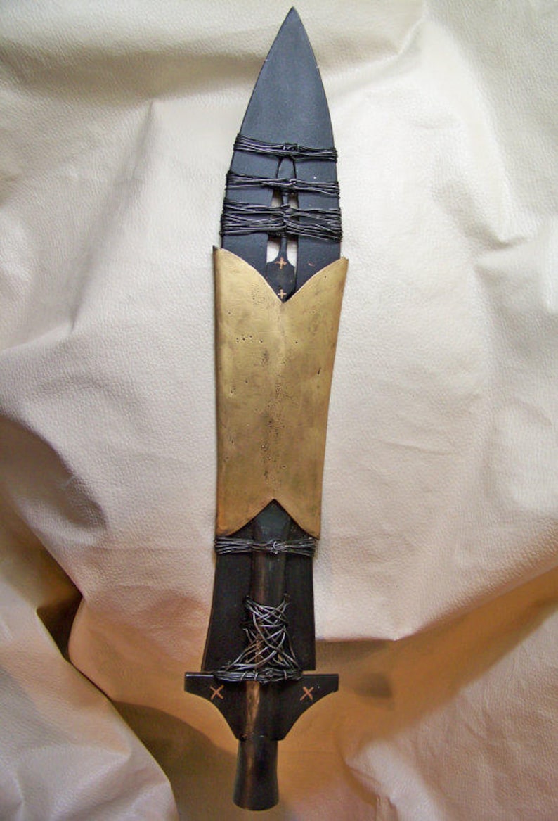 May include: A black and gold spear with a pointed tip and a decorative metal band around the middle. The spear is wrapped in black cord and has a wooden handle.