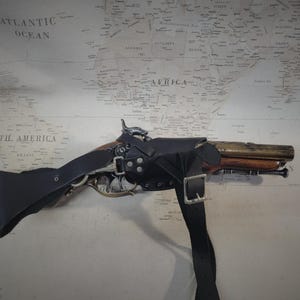 May include: A black leather holster with a brown and silver pistol. The holster has a strap that goes over the shoulder.