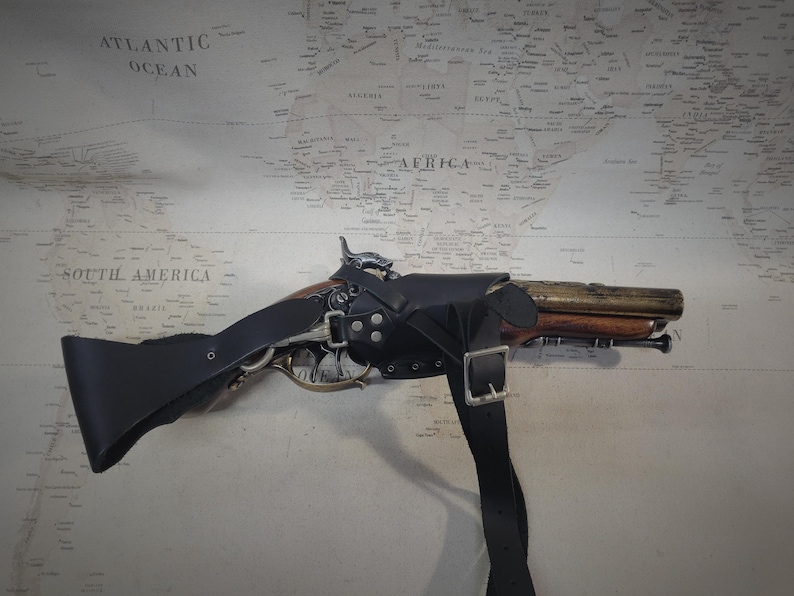 May include: A black leather holster with a brown and silver pistol. The holster has a strap that goes over the shoulder.