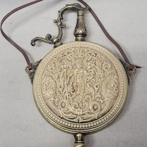 Peut inclure: Une flasque de style antique avec une face circulaire crème ornée d'une sculpture en relief d'une scène mythologique. La flasque a un cadre, une poignée et une base en métal de couleur bronze, avec une sangle en cuir marron.