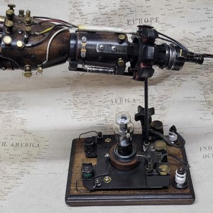 May include: A mechanical prosthetic hand sculpture mounted on a dark wooden base. The hand features dark brown wood, brass joints, and exposed wiring. The base includes a central glass vacuum tube, small metal components, and a vertical support rod.