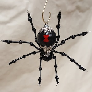 Puede incluir: Adorno de araña de cuentas negras con un reloj de arena rojo en el abdomen y adornos plateados.