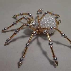 May include: A decorative spider ornament with a body of white pearls and a silver and gold head. The spider's legs are made of gold wire with silver and gold beads. This handcrafted spider is a unique Halloween decoration.