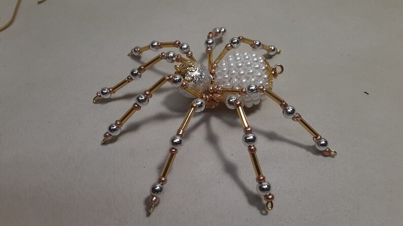 May include: A decorative spider ornament with a body of white pearls and a silver and gold head. The spider's legs are made of gold wire with silver and gold beads. This handcrafted spider is a unique Halloween decoration.