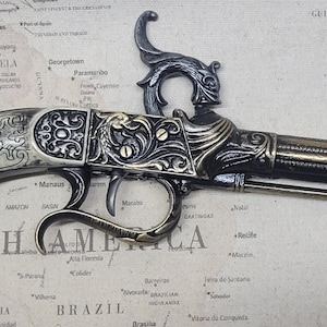 Può includere: Una replica decorativa di pistola a pietra focaia con un'impugnatura color argento decorata con intricati intagli a volute, una canna in metallo scuro e un meccanismo a cane curvo. La superficie metallica presenta dettagliati motivi floreali.