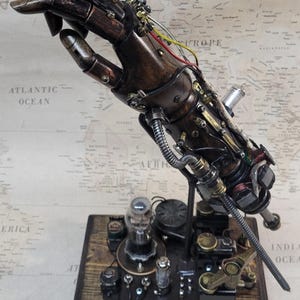 Può includere: Una scultura di mano meccanica in stile steampunk. La mano è realizzata in metallo marrone scuro con accenti in rame, fili e piccoli componenti meccanici. È montata su una base di legno con parti elettroniche, creando un'opera d'arte unica.