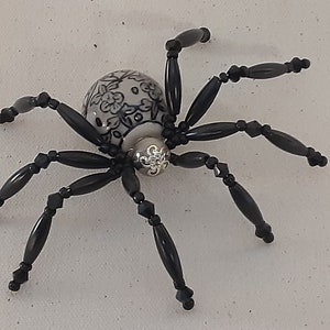 May include: A black beaded spider with a white ceramic body decorated with a black floral pattern. The spider has eight legs and a silver charm in the center of its body.