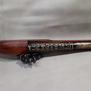 May include: A decorative, antique-style flintlock pistol with a wooden handle and a metal barrel. The barrel is decorated with intricate gold-colored designs.