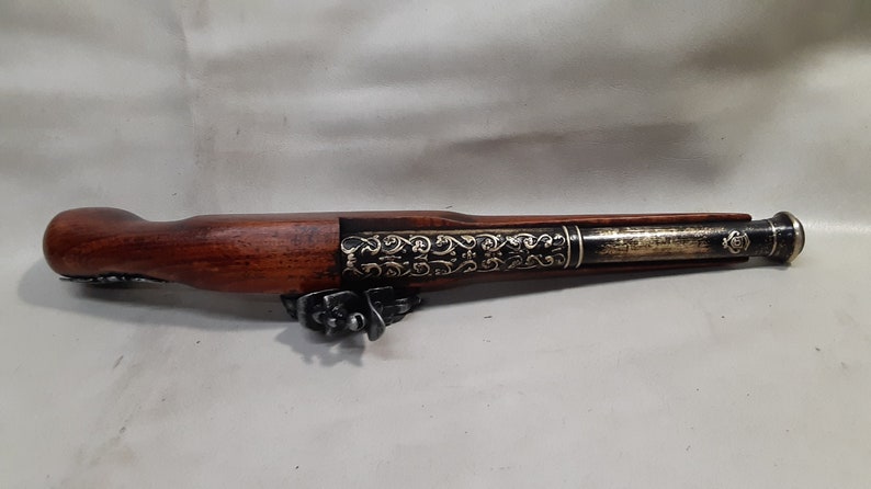 May include: A decorative, antique-style flintlock pistol with a wooden handle and a metal barrel. The barrel is decorated with intricate gold-colored designs.