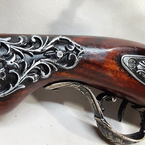 May include: A decorative antique-style pistol with a wooden handle and ornate metal accents. The handle is decorated with a silver and black floral design. The pistol has a silver trigger guard and a silver hammer.