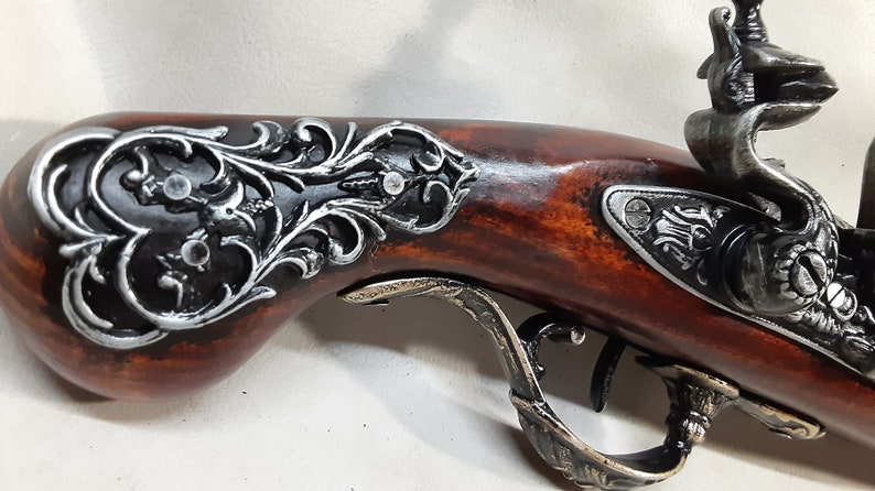 May include: A decorative antique-style pistol with a wooden handle and ornate metal accents. The handle is decorated with a silver and black floral design. The pistol has a silver trigger guard and a silver hammer.
