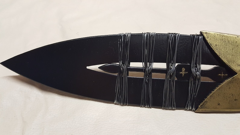 May include: Close-up of a black spearhead with a gold tip. The spearhead is wrapped with black wire.
