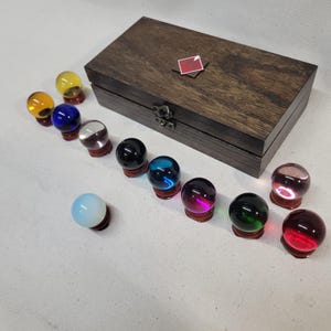 Könnte beinhalten: Eine Sammlung farbiger Glaskugeln auf Holzständern, angeordnet vor einer dunklen Holzkiste. Die Kugeln sind in verschiedenen Farben, darunter Gelb, Blau und Rot. Die Kiste hat ein kleines rotes Etikett oben.