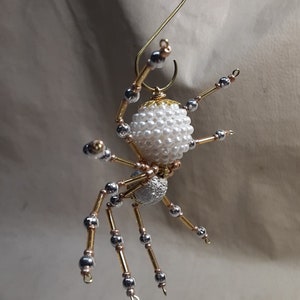 May include: A handmade spider ornament with a white pearl bead body and silver and gold-colored legs. The spider is suspended by a gold wire hook, perfect for holiday or Halloween decor.