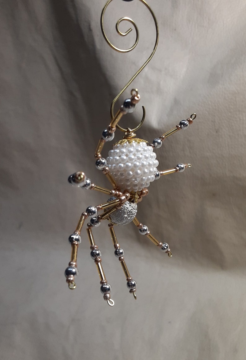 May include: A handmade spider ornament with a white pearl bead body and silver and gold-colored legs. The spider is suspended by a gold wire hook, perfect for holiday or Halloween decor.