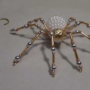 May include: A decorative spider ornament with a pearly white body and gold accents. The spider's legs are made of gold and silver beads. A gold-colored wire spiral is to the left of the spider.