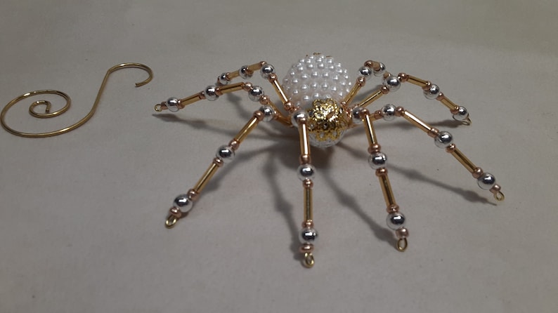 May include: A decorative spider ornament with a pearly white body and gold accents. The spider's legs are made of gold and silver beads. A gold-colored wire spiral is to the left of the spider.