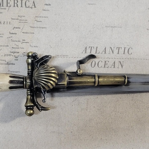 May include: An antique letter opener with a decorative handle and a silver-colored blade. The handle features a cream-colored grip with ornate gold-colored metalwork. The blade is long and pointed, with a slight curve. The opener is lying on a map.